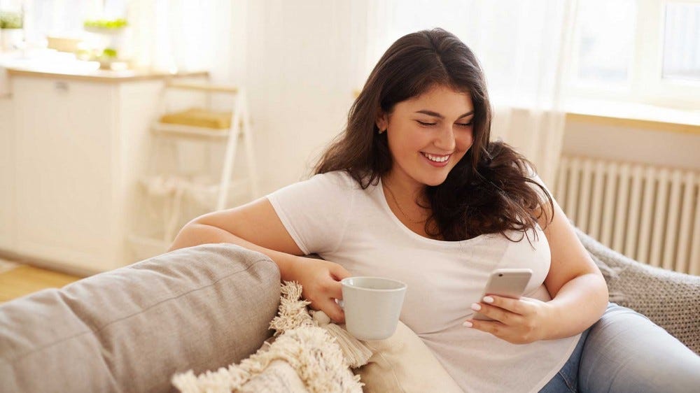 O tânără care stă pe o canapea și bea cafea, zâmbind în timp ce folosește o aplicație fericire pe smartphone.