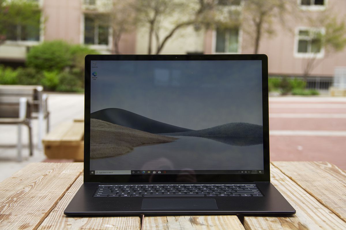 Laptopul Surface 4 de 15 inci se deschide pe o masă de picnic cu o clădire de apartamente roz în fundal.  Ecranul afișează dune de nisip cu un cer albastru.