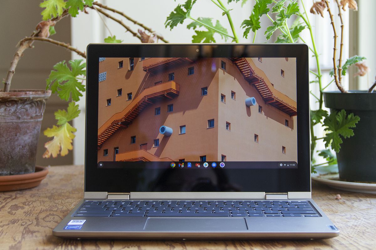 Chromebookul Lenovo Ideapad Flex 3 stă ​​deschis pe o masă în fața a două plante de apartament.  Ecranul afișează ferestrele superioare ale unei clădiri mari.