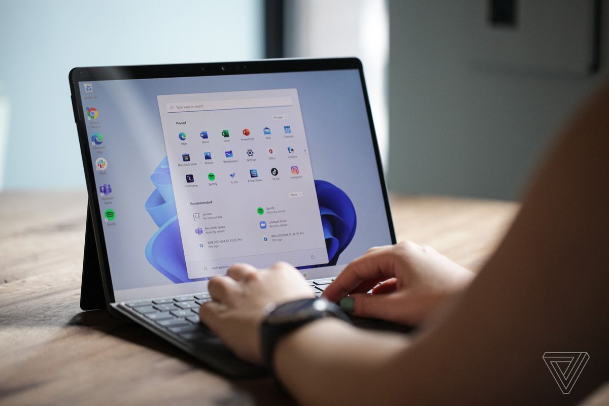 Un utilizator tastează pe Surface Pro 8 din spate.  Ecranul afișează meniul de pornire Windows 11 pe un fundal alb și albastru.