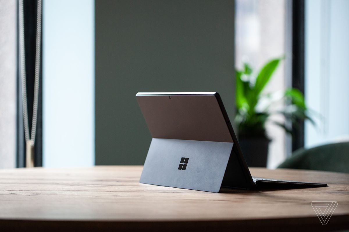 Surface Pro 8 de pe o masă văzută din spate spre dreapta, deschisă, cu tastatura semnată atașată și suportul afară.  În fundal este un perete verde și o plantă în ghiveci.